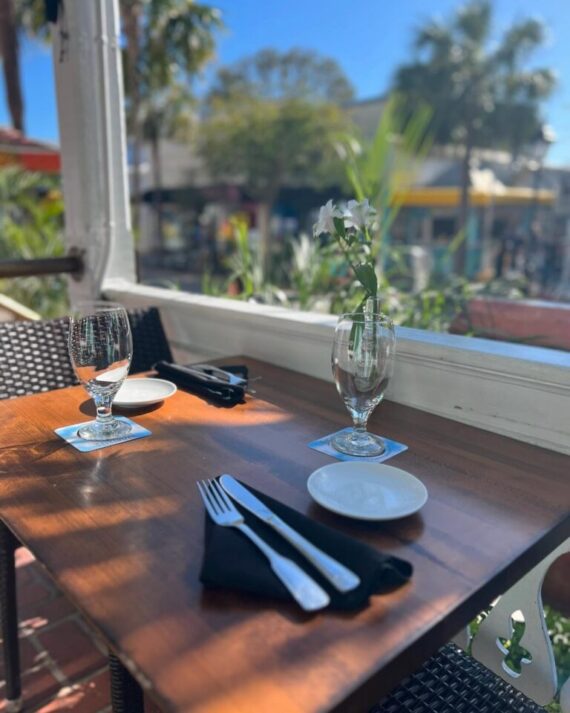 A wooden outdoor restaurant table set for two with glasses, plates, cutlery, and a single flower in a vase, surrounded by greenery and a sunny, blurred street scene in the background.