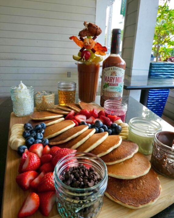 A wooden board with pancakes, strawberries, blueberries, banana slices, jars of chocolate chips, yogurt, and whipped cream. In the background is a Bloody Mary with garnishes and a bottle of Bloody Mary mix.