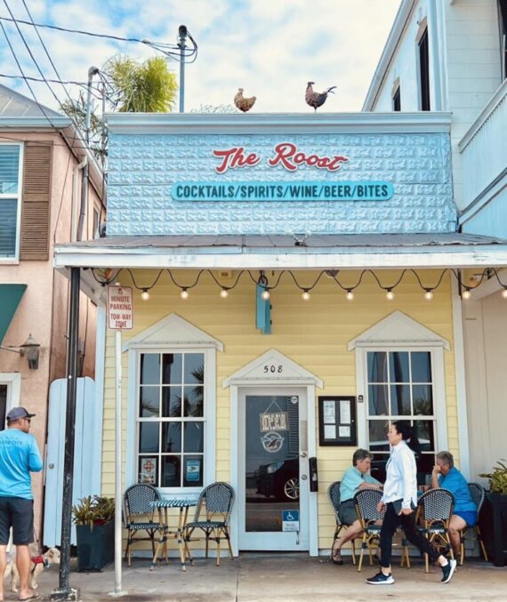 A small yellow café called The Roost with people sitting at outdoor tables, a man and woman walking by, and two roosters on the roof. The sign advertises cocktails, spirits, wine, beer, and bites.