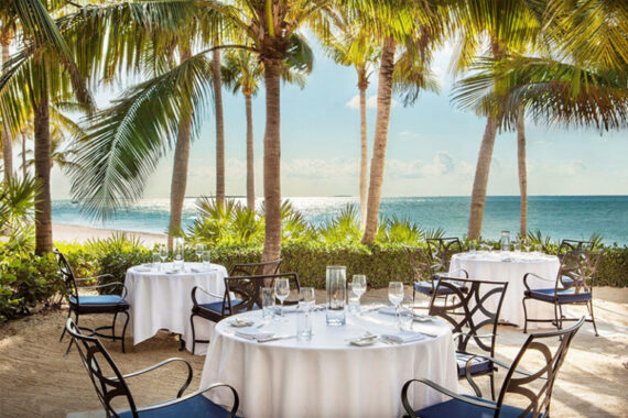Outdoor dining area with round tables covered in white cloths, set with glassware and cutlery, surrounded by palm trees, overlooking a sunny beach and the ocean.