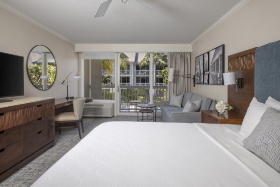 Modern hotel room with a large bed, wooden furniture, desk with chair, sofa, round mirror, flat-screen TV, and sliding glass doors leading to a balcony with a view of palm trees outside.