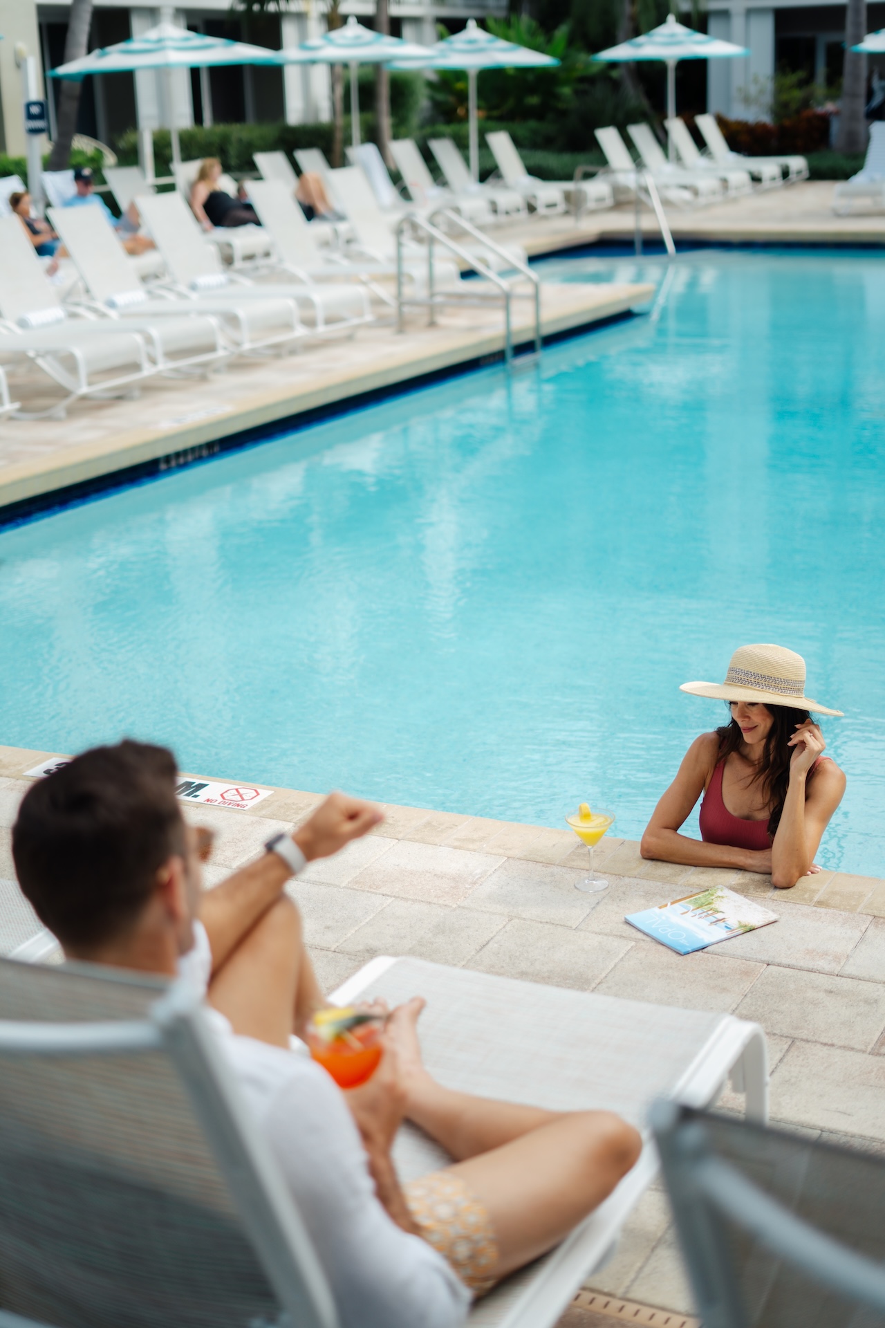 A woman in a sunhat relaxes in a pool and talks on the phone while a man lounges nearby with a drink. There are lounge chairs and umbrellas by the poolside in the background.