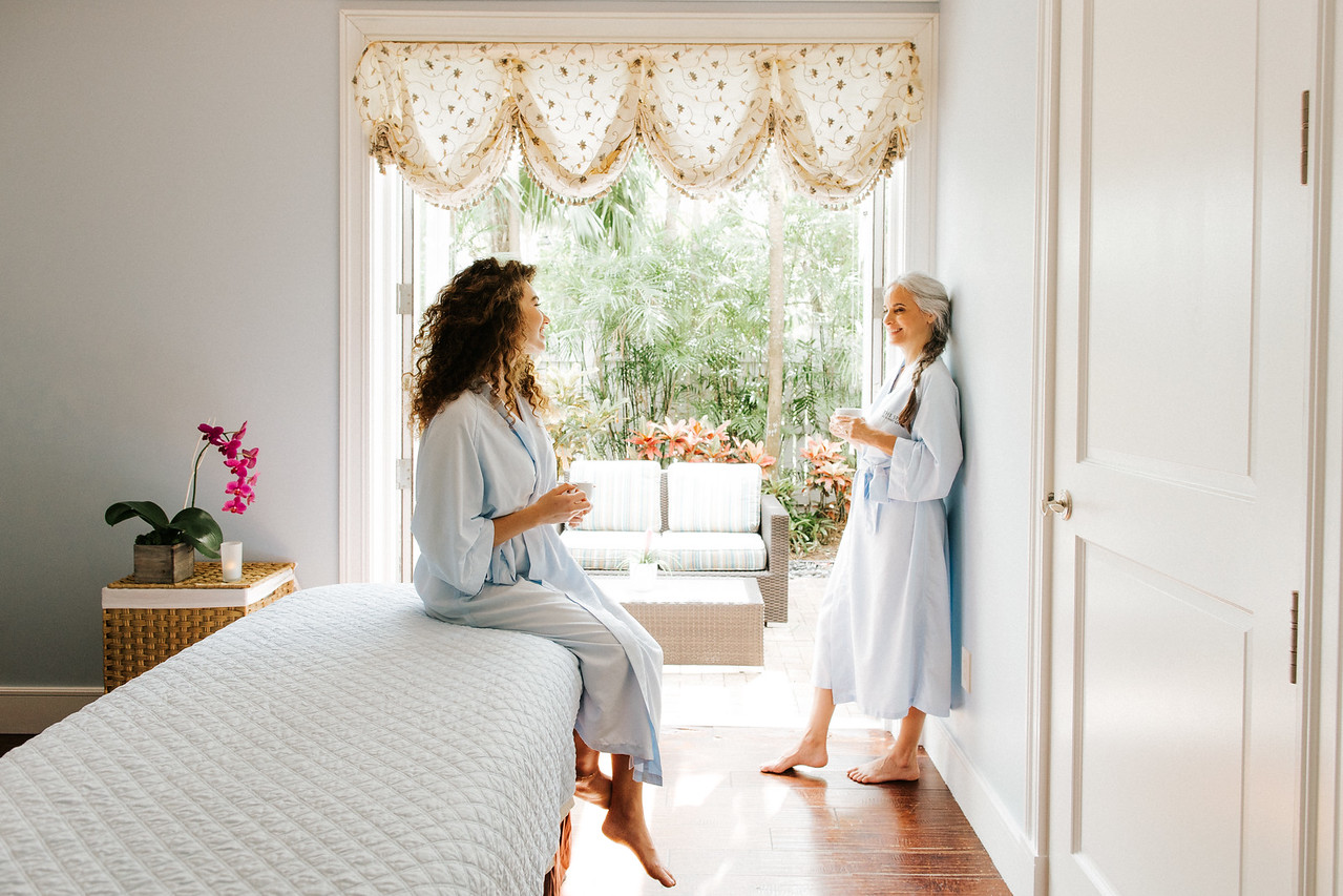 Two women in light robes chat and smile in a bright, cozy bedroom with an orchid and garden view. One sits on the bed, the other leans against the wall, both holding mugs.
