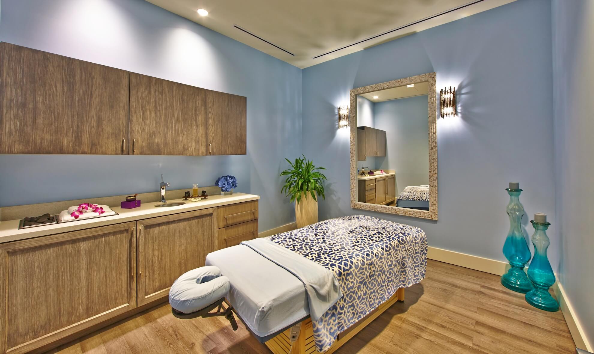 A serene spa room with a massage table covered in a patterned blue and white sheet. Wooden cabinets line the left wall, topped with flowers and decor. A large mirror hangs on the right, with two tall blue glass vases nearby. Soft lighting creates a calming ambiance.