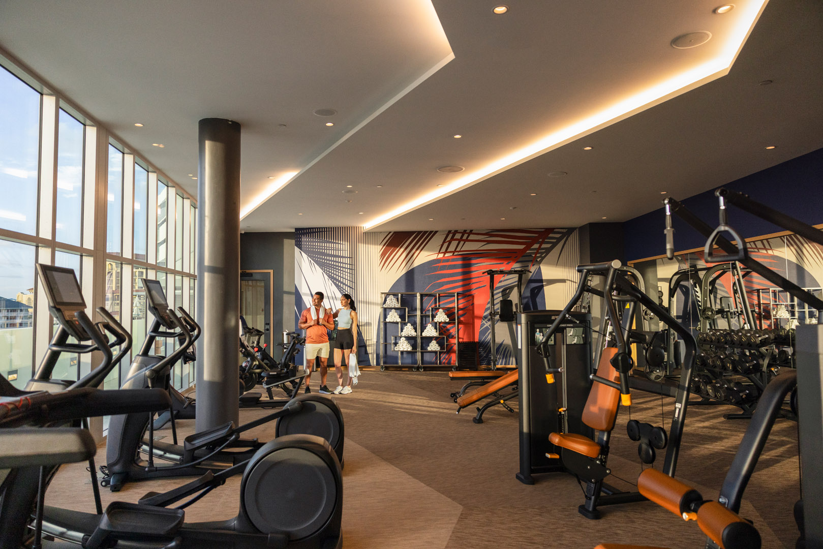 Modern gym with cardio and weight machines, large windows showing a city view reminiscent of Clearwater Beach, a mural on the back wall, and two people standing and talking near the center.