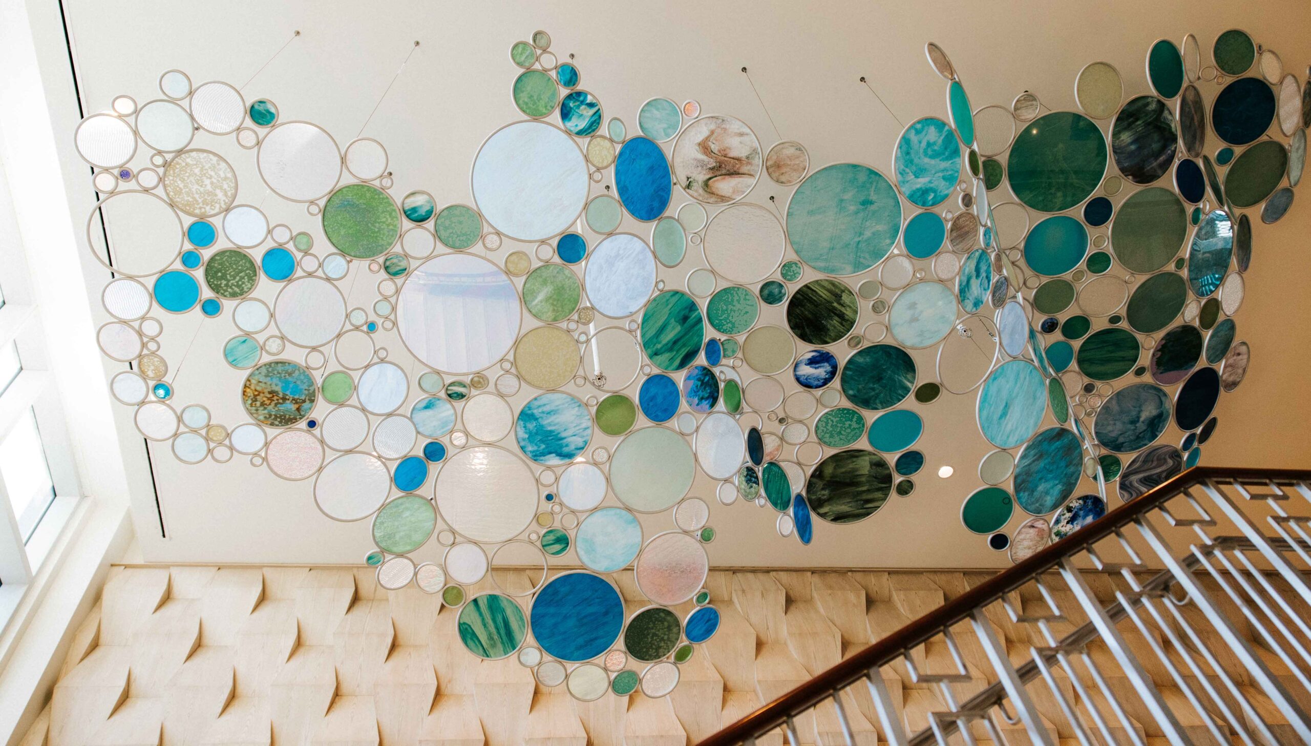 Hanging art installation of variously sized circular glass pieces in blue, green, and white tones, reminiscent of the tranquil hues found at Clearwater Beach, suspended from the ceiling above a staircase with a geometric railing and beige textured wall.