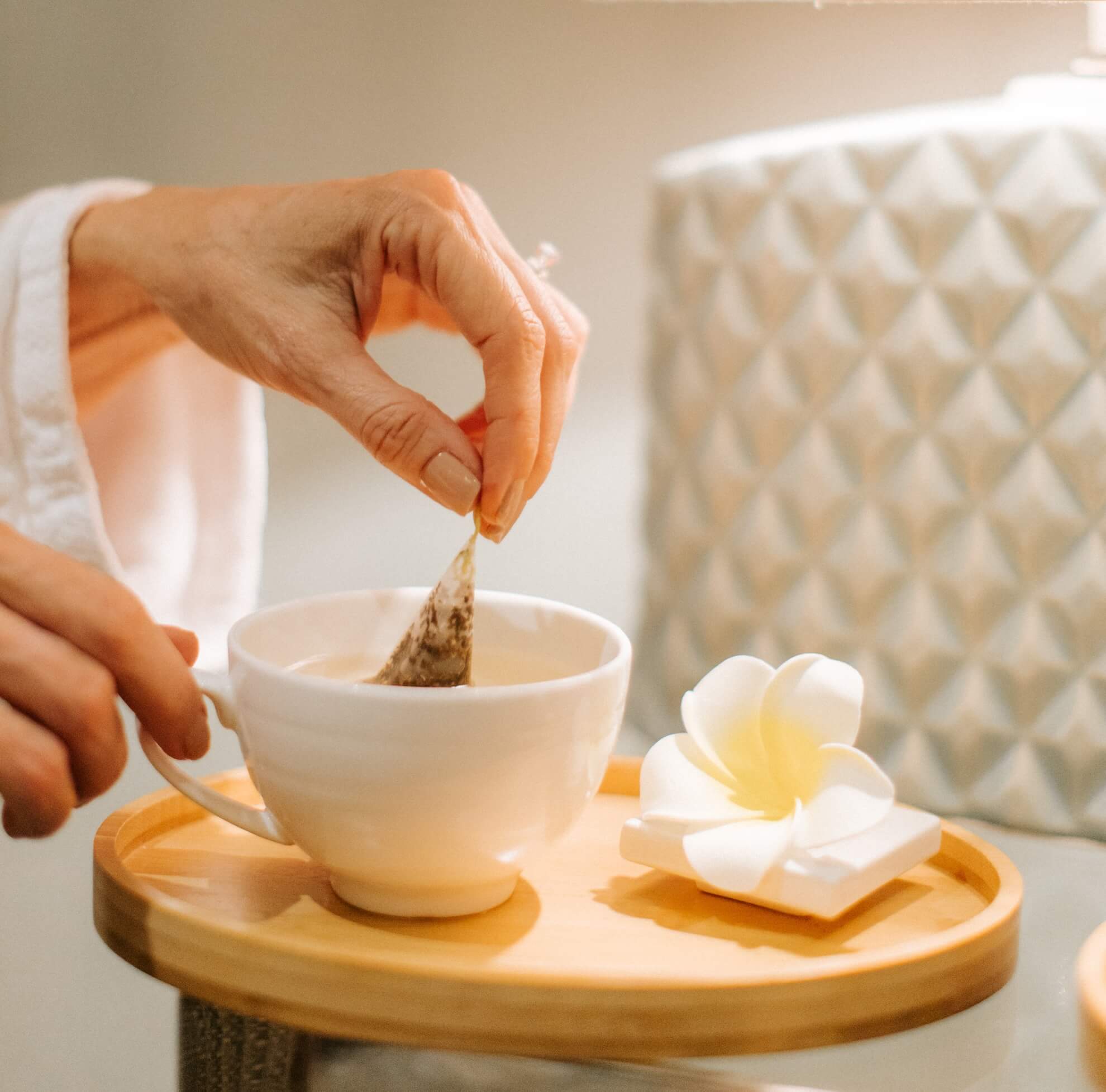 Close-up of a person in a robe dipping a teabag into a white cup on a wooden tray. A plumeria-shaped soap or decoration is next to the cup. The background features a textured, diamond-patterned lamp.