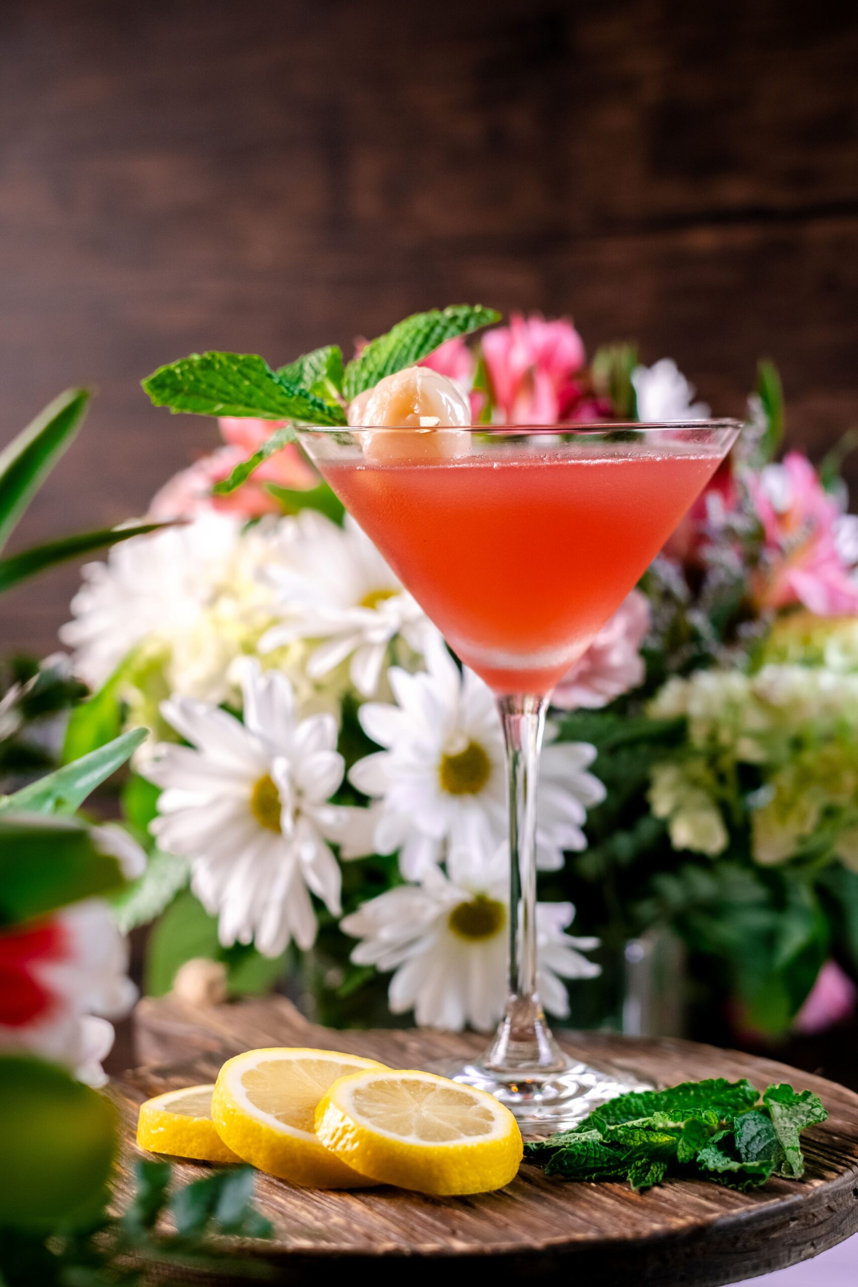 A pink cocktail in a martini glass garnished with a lychee and mint sprig, set on a wooden surface with lemon slices and fresh mint. Colorful flowers with white daisies are blurred in the background.