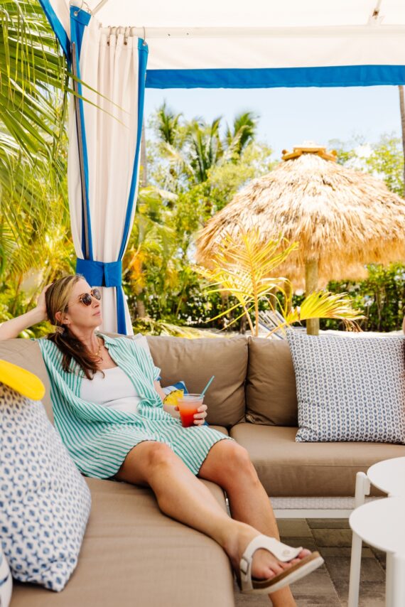 A woman in sunglasses relaxes on an outdoor couch under a cabana, holding a colorful drink. She is surrounded by tropical plants and a thatched umbrella in a sunny, resort-like setting.