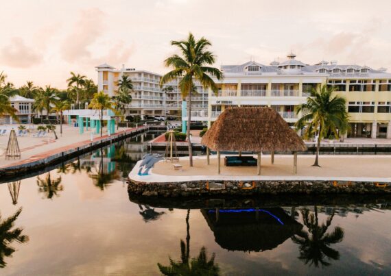 A waterfront resort with palm trees, a thatched-roof hut, and modern buildings reflects in calm water at sunset, creating a serene tropical atmosphere.