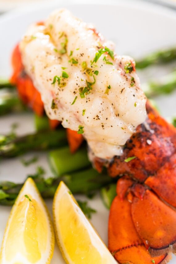 A cooked lobster tail topped with herbs sits on asparagus spears, with two lemon wedges on the side. The dish is served on a white plate.