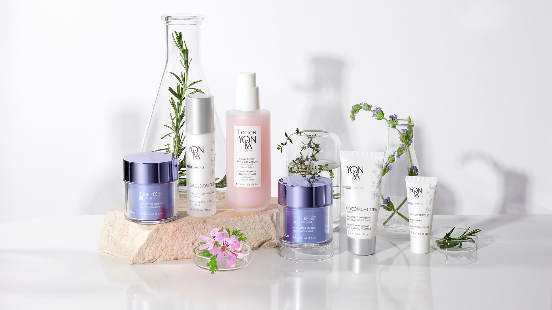 A display of Yon-Ka skincare products arranged on a rock with flowers and sprigs of rosemary, set against a white background with soft shadows.
