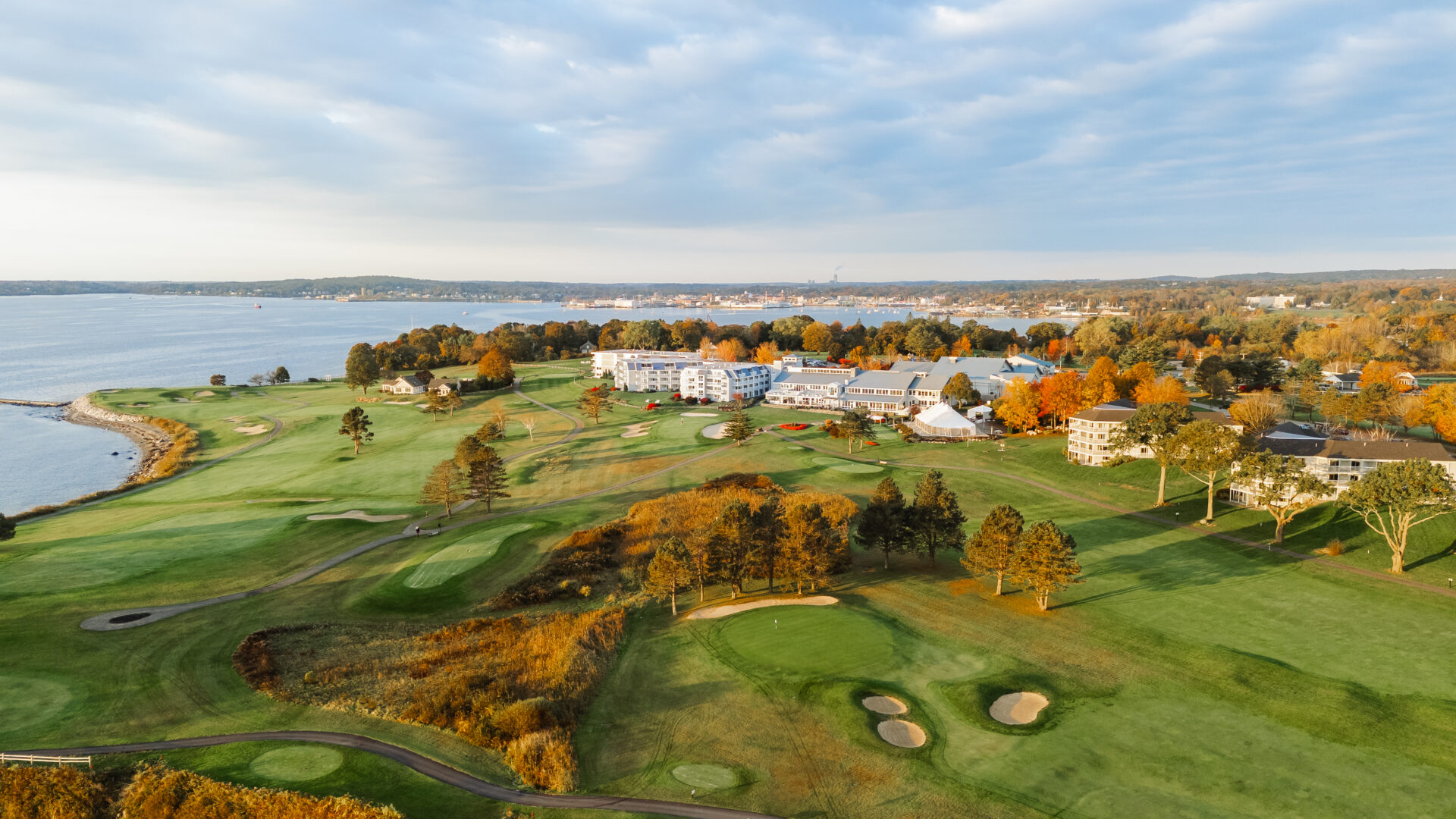 Aerial view of a scenic golf course next to a body of water. The landscape features lush green fairways, sand traps, and autumn-colored trees. A resort with several buildings is visible in the background under a partly cloudy sky.