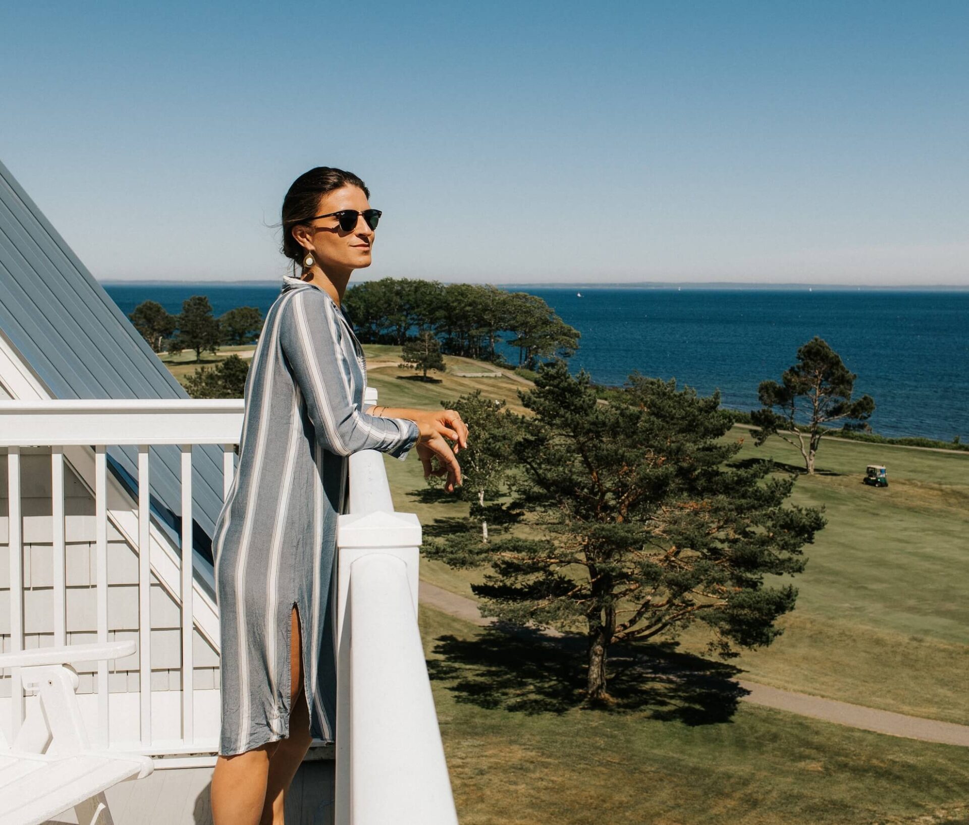 A woman wearing sunglasses and a striped dress stands on a balcony overlooking a scenic landscape with trees, grass, and a view of the ocean on a clear, sunny day.