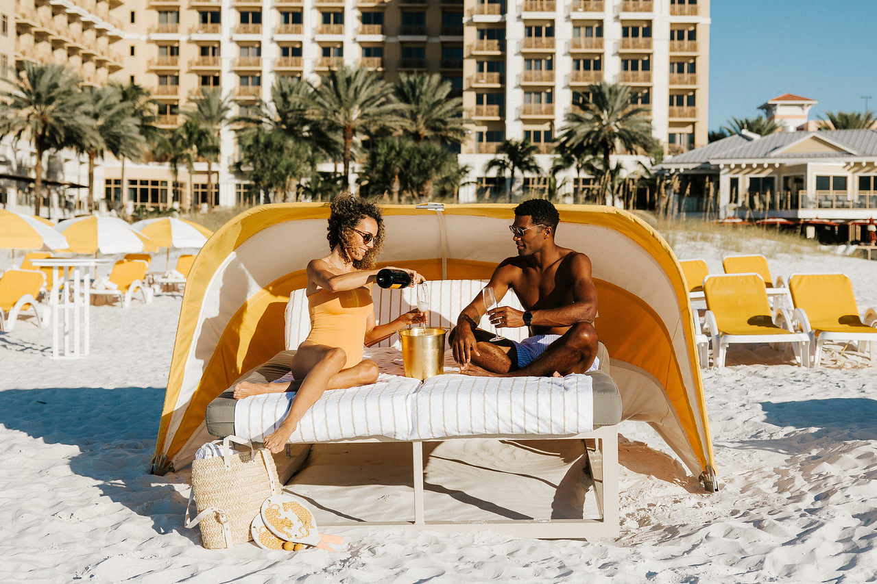 A couple relaxes on a beach canopy bed, dressed in swimsuits. The woman pours a drink from a bottle into a glass on a table between them. Background shows palm trees, a building, and more lounging areas with yellow umbrellas.