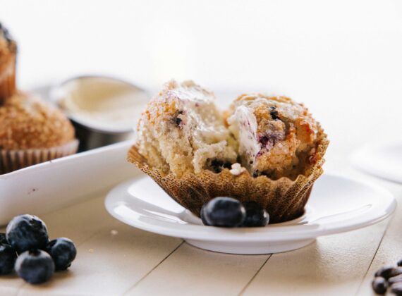 A split blueberry muffin on a white plate, revealing its soft, crumbly interior. Fresh blueberries are scattered around the table, and theres a blurred muffin tin in the background.