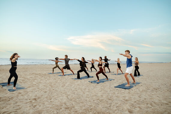 A group of people practice yoga on mats on a sandy beach at sunset, all standing in a warrior pose and facing the ocean under a clear sky.