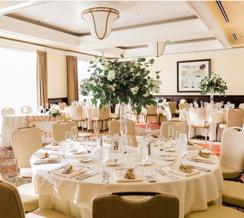 Elegant banquet hall set for an event, with round tables covered in white linens, arranged chairs, neatly set tableware, and tall green floral centerpieces. Warm natural light streams in through large windows.