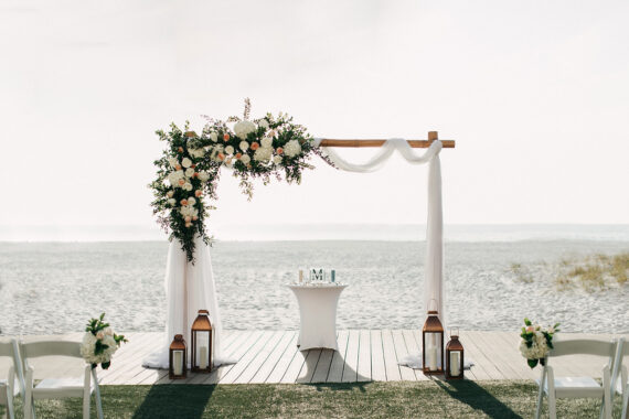 A wooden wedding arch draped in white fabric and adorned with white and blush flowers stands on a beach. Lanterns and flower arrangements line the aisle, and the ocean is visible in the background.