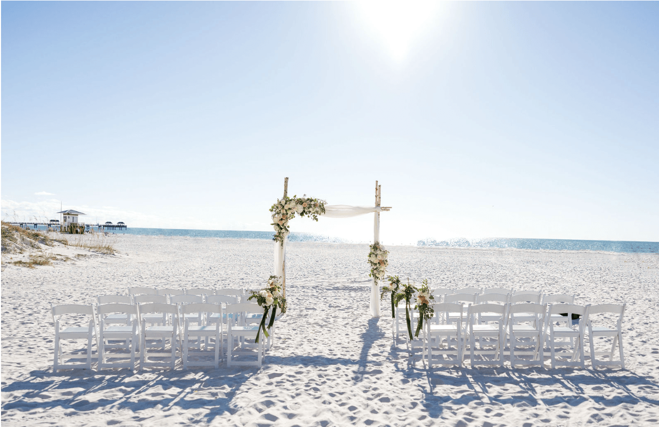A beach wedding setup with white chairs arranged in rows facing a floral archway on the sand, overlooking the ocean under a bright, clear sky.