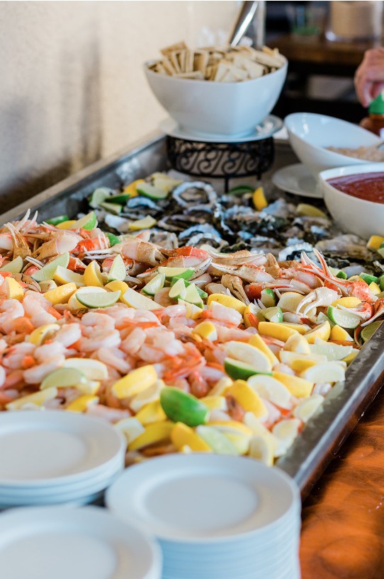 A buffet table features assorted seafood, including shrimp and crab, garnished with sliced lemon and lime. Plates are stacked in the foreground, with bowls of crackers and dipping sauce in the background.