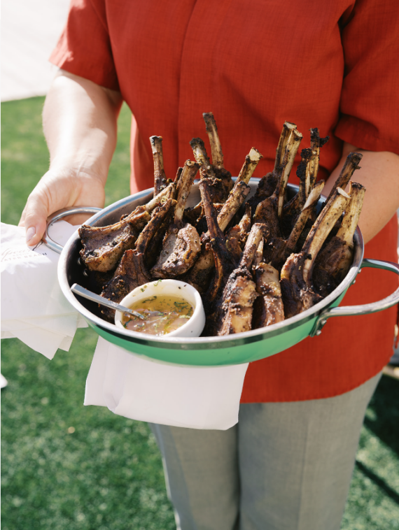 A person in a red shirt holds a green metal bowl filled with grilled lamb chops, accompanied by a small dish of dipping sauce. The scene is outdoors on green grass.