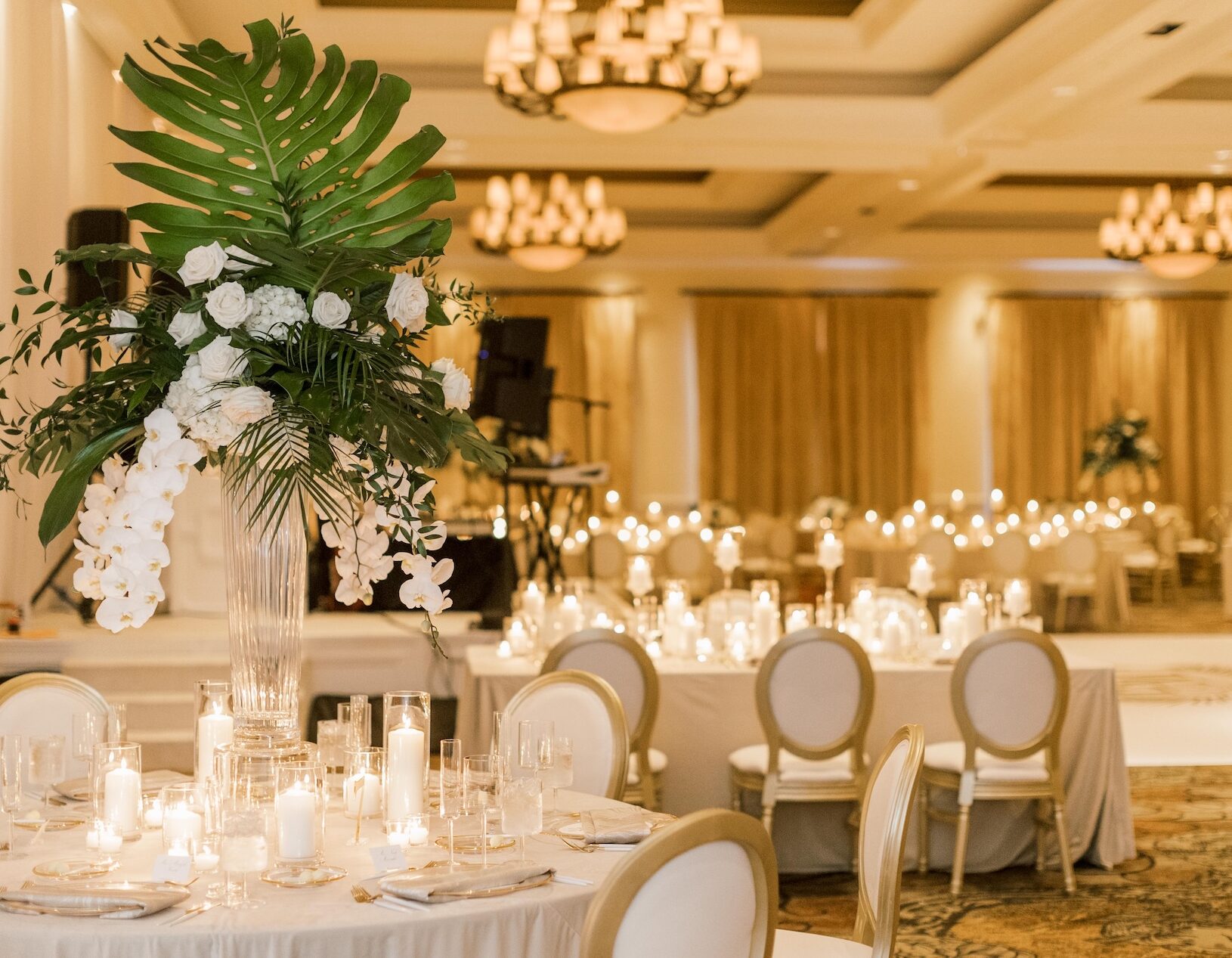 Elegant banquet hall set for an event with round tables adorned with white linens, tall floral centerpieces, and numerous lit candles; chandeliers hang from the ceiling, creating a warm, inviting ambiance.