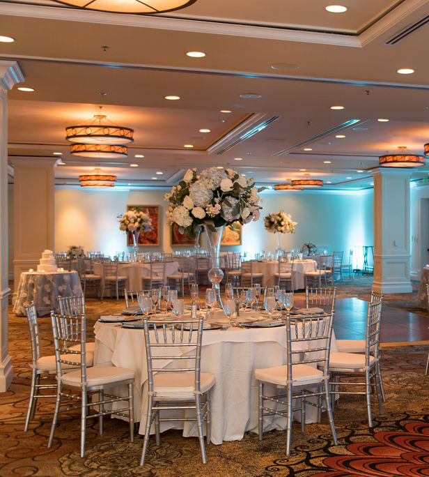 A spacious, elegant banquet hall set for a formal event, with round tables covered in white cloths, silver chairs, and large white floral centerpieces under warm, decorative ceiling lights.