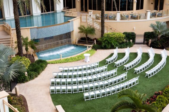 Rows of white chairs are arranged in a semicircle on a lawn, facing a small round table, with a pool, waterfall feature, and lush tropical plants in the background.