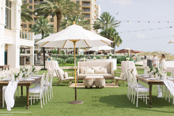 Outdoor wedding reception setup on grass with white tables, chairs, a sofa, and umbrellas. Palm trees, string lights, and a beachfront hotel are visible in the background on a sunny day.