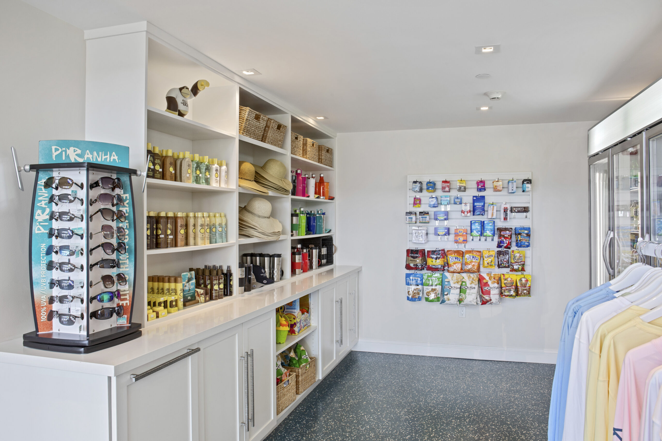 A bright, modern store with white shelves stocked with sunscreen, hats, snacks, and toiletries. Sunglasses are displayed on a rotating stand, and colorful shirts hang on a rack to the right.
