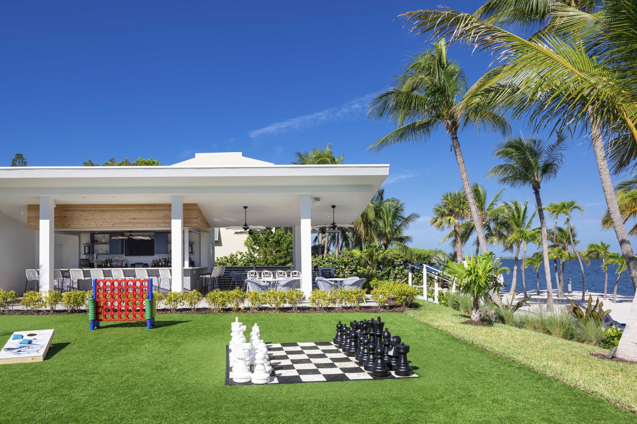 A tropical outdoor lounge with a bar, seating, palm trees, giant chess set, Connect 4, and cornhole on a green lawn, overlooking the ocean under a clear blue sky.