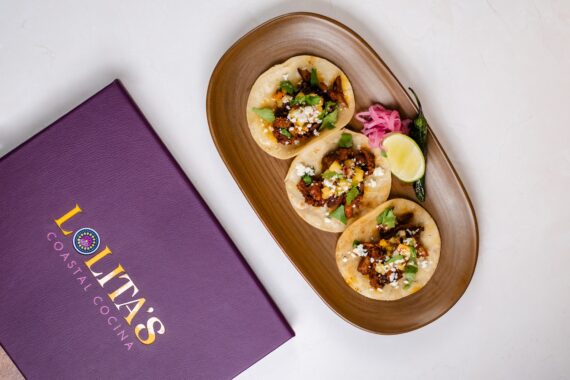 Three street-style tacos topped with meat, cheese, greens, and avocado are served on a brown plate with lime, pickled onions, and a roasted jalapeño, next to a purple menu labeled Lolitas Coastal Cocina.