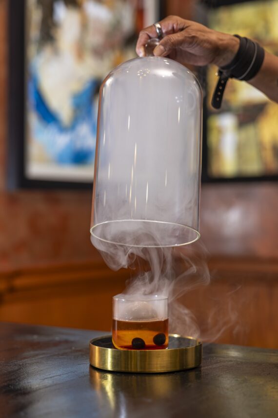 A hand lifts a glass cloche, releasing smoke from a cocktail garnished with two dark berries in a glass on a gold coaster, set on a dark table with an abstract art background.