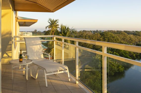 A sunny balcony with a lounge chair and a small table holding a bottle of wine and two glasses overlooks green trees, a river, and distant buildings under a clear sky.