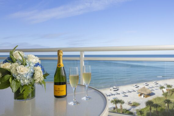 A table on a balcony overlooking a beach with a bouquet of blue and white flowers, a bottle of Veuve Clicquot champagne, and two filled champagne glasses, with the ocean and loungers in the background.