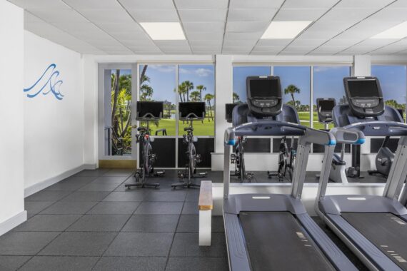 A modern gym with treadmills and stationary bikes facing large windows, offering a view of palm trees and a grassy outdoor area. The room has white walls, a black floor, and a blue wave design on one wall.