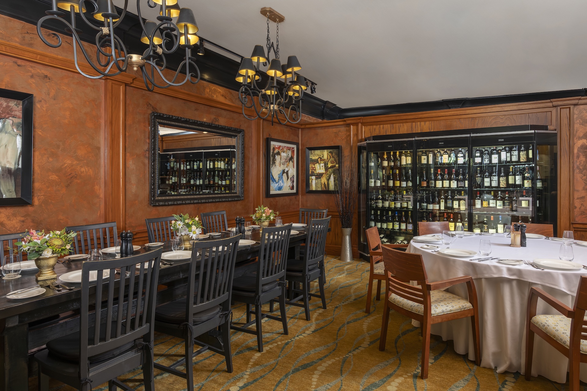 Private dining room with orange-brown walls, chandeliers, a wine display, two dining tables—one rectangular with black chairs and one round with beige chairs—both set for meals with flowers and tableware.