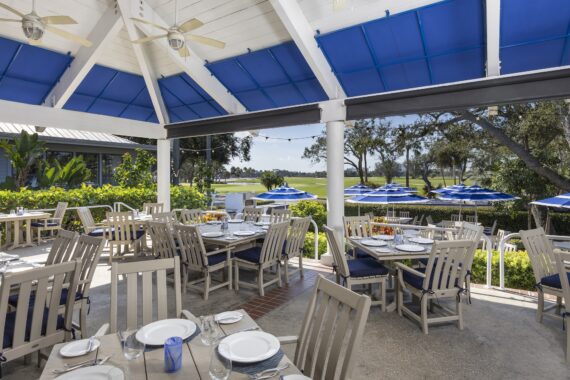Outdoor dining area with wooden tables and chairs set for a meal, under a white pavilion with blue accents. Blue umbrellas and lush greenery surround the space, with a scenic view of trees and open fields in the background.