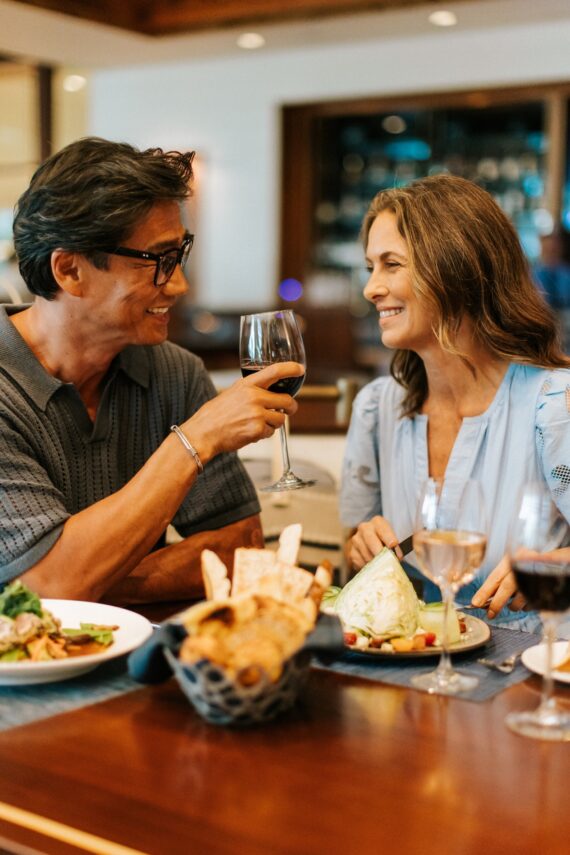 A man and woman sit at a restaurant table, smiling at each other while holding glasses of wine. Plates of food and bread are in front of them, creating a warm, cheerful dining atmosphere.