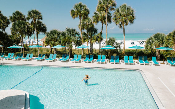 Person swimming in a large outdoor pool surrounded by turquoise lounge chairs and umbrellas at The Resort at Longboat Key Club.