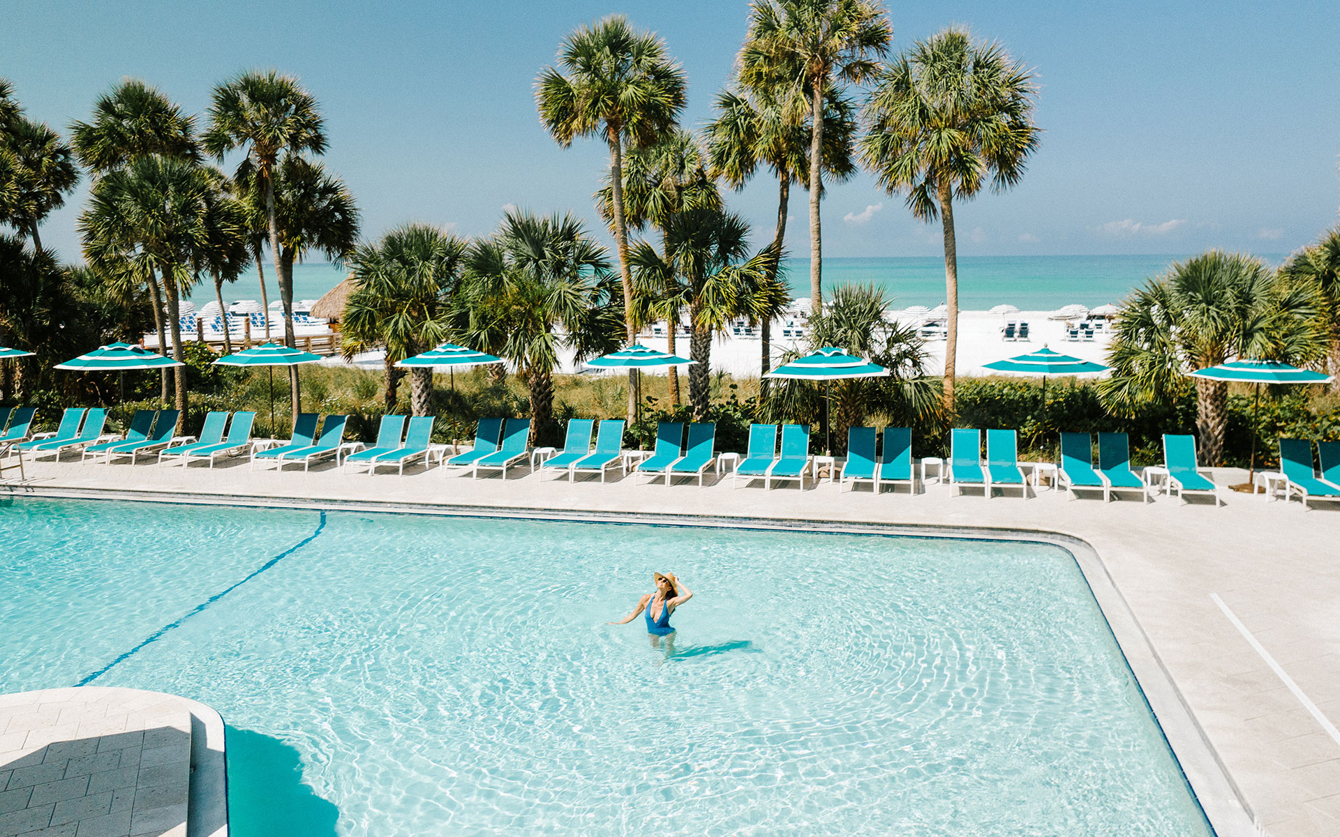 Person swimming in a large outdoor pool surrounded by turquoise lounge chairs and umbrellas at The Resort at Longboat Key Club.