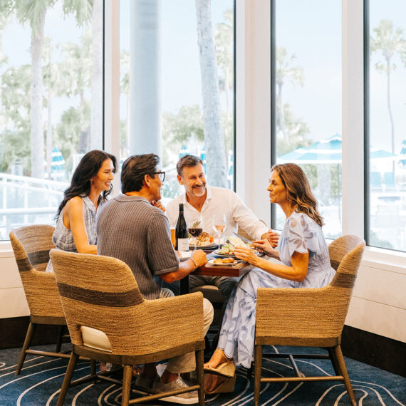 Four adults sit around a table in a bright, modern restaurant, enjoying food and drinks, smiling and talking. Large windows reveal palm trees and an outdoor area with umbrellas in the background.