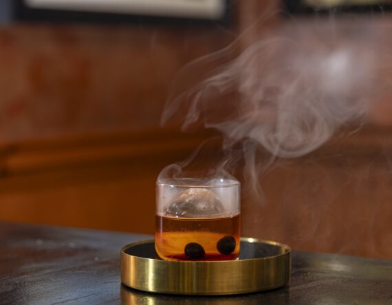 A glass of amber cocktail with ice and two dark cherries sits on a round brass tray. Wisps of smoke rise from the drink, set on a dark table with a warm, blurred background.