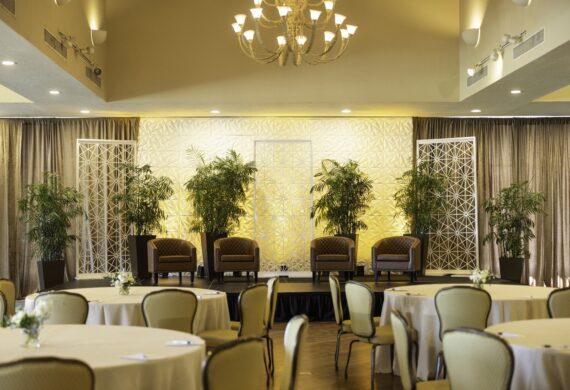 A conference room with round tables and chairs faces a stage with four armchairs, surrounded by potted plants and decorative screens, under warm lighting and a chandelier.