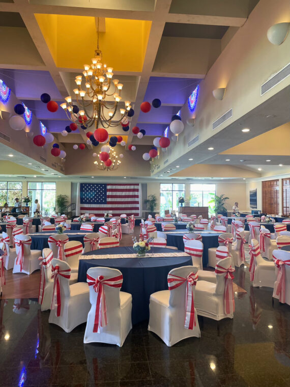 A banquet hall decorated with red, white, and blue balloons and ribbons, featuring round tables and chairs with patriotic covers. An American flag hangs on the back wall, with chandeliers and natural light filling the room.