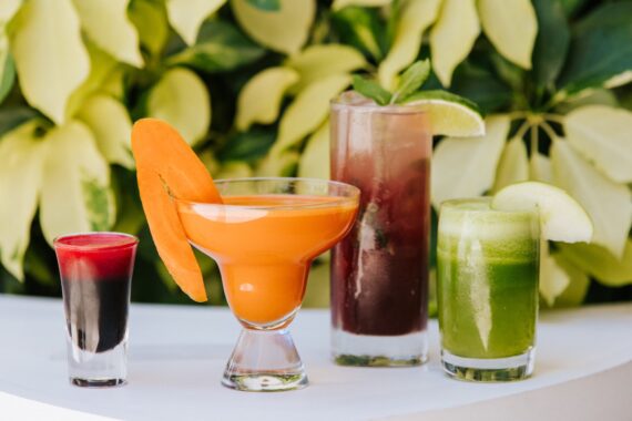 Four colorful drinks on a white surface: a red shot, an orange drink in a wide glass with a carrot slice, a tall purple drink with mint and lime, and a green juice with an apple slice. Pale leafy background.