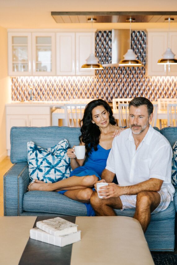 A man and woman sitting on a blue sofa holding mugs in a kitchen.