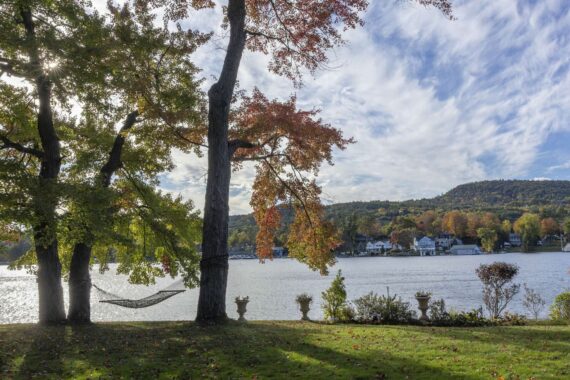 A hammock hangs between two trees on a grassy lawn by a lake, with vibrant autumn foliage, potted plants on a stone railing, and hills and houses visible across the water under a partly cloudy sky.