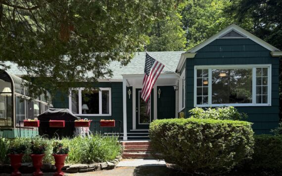 A green house with white trim, a front porch, and an American flag above the steps. Red planters with flowers line the walkway, and a grill is on the left patio. Trees and bushes surround the house on a sunny day.